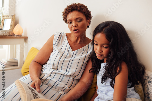 Granny read to her granddaughter in the living room. Kid and grandma are sitting together on a sofa.