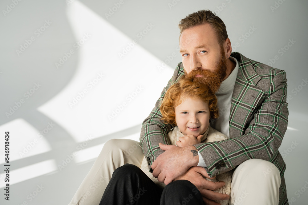 Bearded father hugging redhead son in knitted jumper on grey background with sunlight Bearded father hugging redhead son in knitted jumper on grey background with sunlight