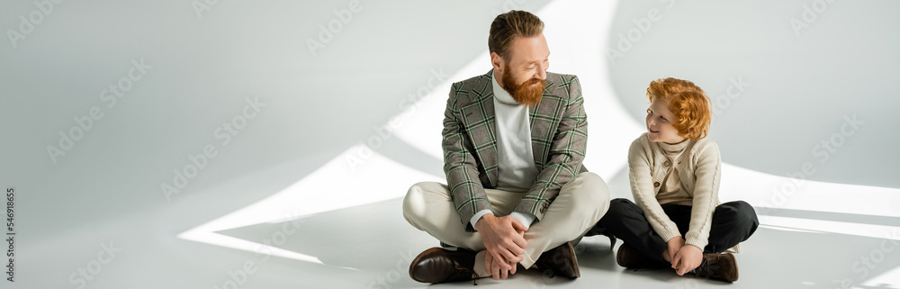 Bearded man in checkered jacket looking at redhead son on grey background with light, banner