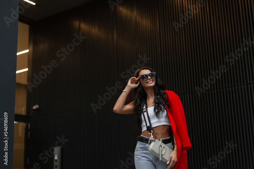 Fashion beautiful business girl in stylish casual clothes with top, jeans, red jacket and bag walking in the city and wearing stylish sunglasses near a modern building