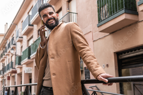 Bearded young man in turtleneck sweater and jacket looking to the side smiling while making a phone call.