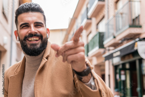 Tableau sur toile Young man with a beard, turtleneck sweater and jacket using his wireless headphones while pointing his index finger at the person he just met