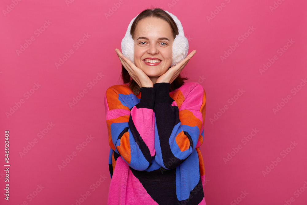 Image of smiling attractive young adult woman wearing winter earmuffs and warm jumper, looking at camera, keeps palms under chin, has cute and charming look, stands isolated over pink background. Image of smiling attractive young adult woman wearing winter earmuffs and warm jumper, looking at camera, keeps palms under chin, has cute and charming look, stands isolated over pink background.