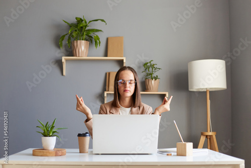 Calm businesswoman taking break for meditation, doing yoga exercise at workplace, mindful attractive female worker feeling zen, no stress free and harmony relaxing at work, sitting in front of laptop.