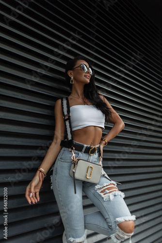 Beautiful stylish fashion girl with a smile with a sexy slim tanned body in fashionable summer clothes with a top, ripped jeans and a handbag stands near a black metal wall on the street