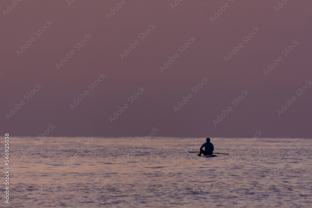 Unrecognizable man sitting on a paddle board in the sea while appreciating the sunrise