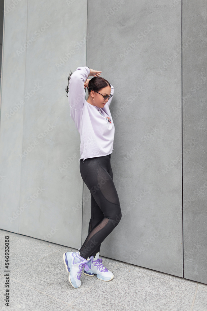 Stylish beautiful plump girl in fashionable sportswear with a purple oversized  sweatshirt and black leggings with trendy sneakers fix her hair near a gray  concrete wall in the city Stock Photo