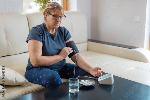 Senior woman checking blood pressure level at home, older female suffering from high blood pressure sitting at a couch and using a pulsometer, tonometer