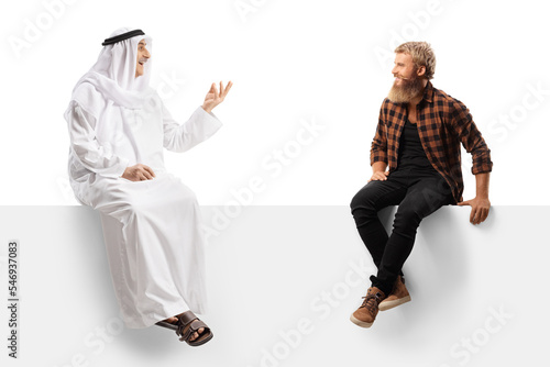 Arab man in an ethnic robe sitting on a blank panel and talking to a casual young man