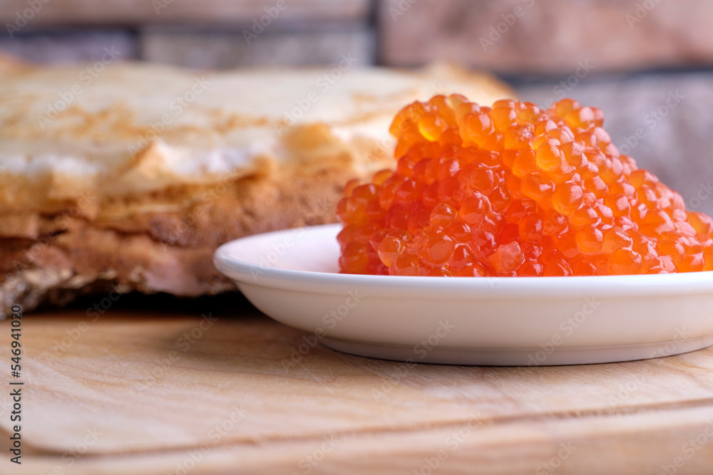 red caviar in a white porcelain bowl next to a stack of thin flour pancakes stands on a wooden stand red caviar in a white porcelain bowl next to a stack of thin flour pancakes stands on a wooden stand