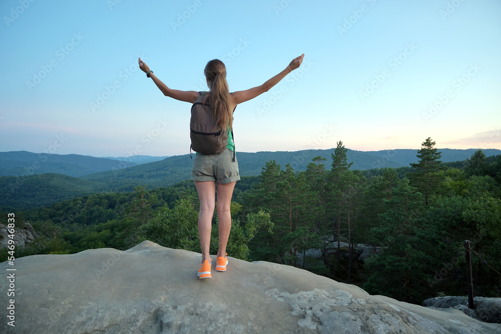 Hiker woman standing on mountain footpath raising up hands enjoying evening nature during travel on wilderness trail. Lonely female traveler traversing high hilltop route. Healthy lifestyle concept Hiker woman standing on mountain footpath raising up hands enjoying evening nature during travel on wilderness trail. Lonely female traveler traversing high hilltop route. Healthy lifestyle concept