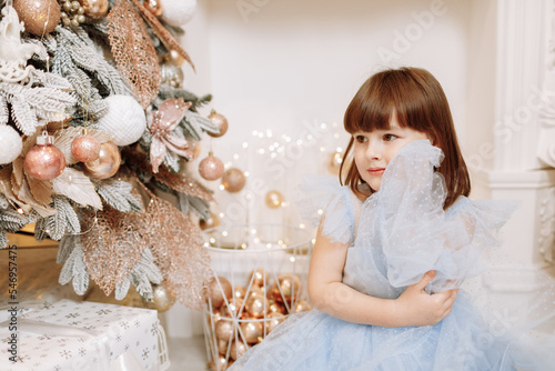 Adorable smiling little girl in festive princess blue dress is looking in camera in cozy room decorated lighting garlands. Happy family holidays, New year. Cozy warm winter evening at home.