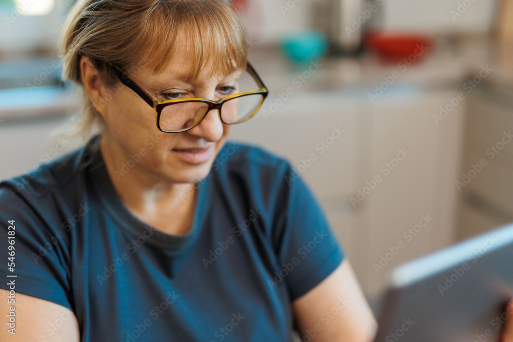 Happy middle aged 50 years old woman using digital tablet sitting in kitchen at home. Mature older lady wearing glasses holding pad computer reading e book, shopping online, making video call. Happy middle aged 50 years old woman using digital tablet sitting in kitchen at home. Mature older lady wearing glasses holding pad computer reading e book, shopping online, making video call.
