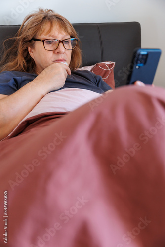 Close-up photo of a sick woman at home, has a bad runny nose and cold, lies in bed covered with a blanket, calls a doctor for advice