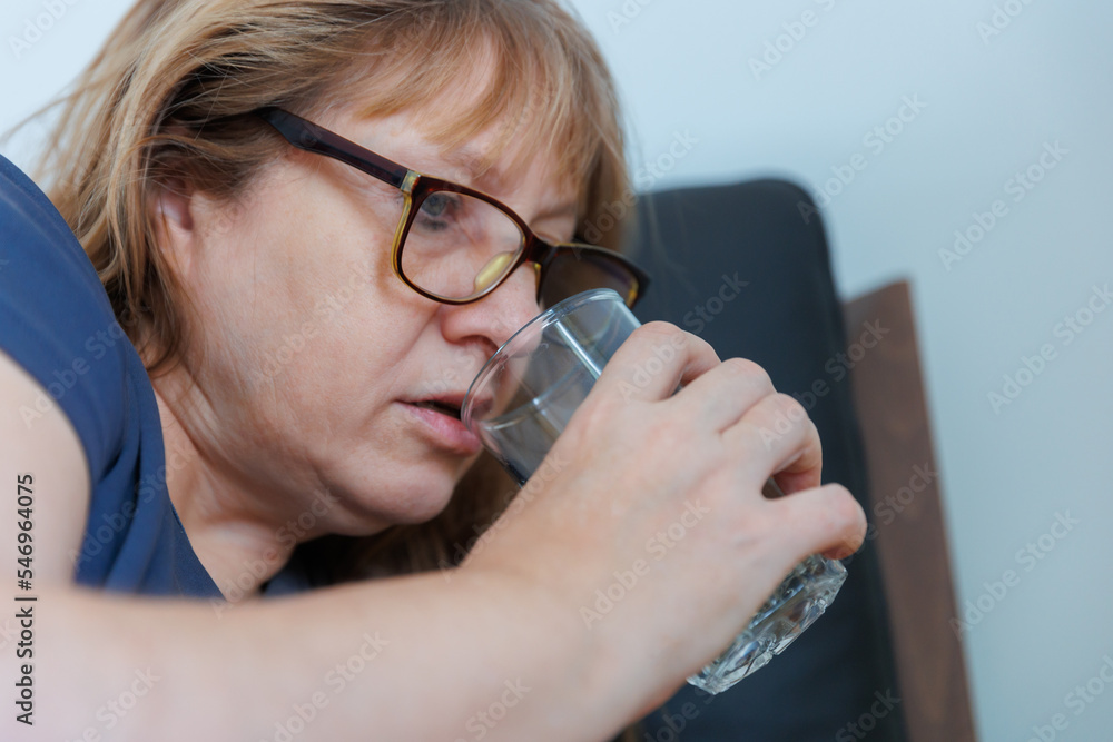 Sick old woman lying in bed at home feel sick suffer from flu or cold cure with prescribed medications. Woman drinking from a glass
