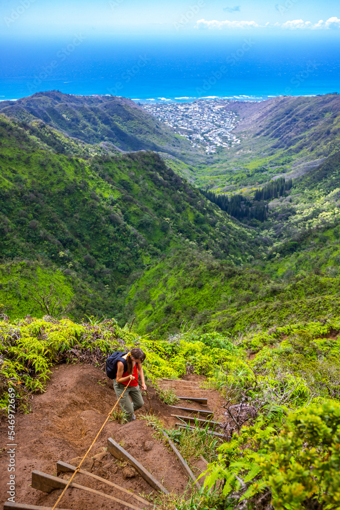 Fototapeta premium hiker girl climbs steep trail on oahu with honolulu skyline in background on wiliwilinui ridge trail; hiking on green mountains in hawaii, holiday in hawaii