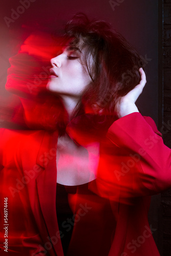 Closeup beauty portrait of young woman with makeup and hairstyle, wear red suit, with closed eyes, holding hand in hair, red neon studio light.