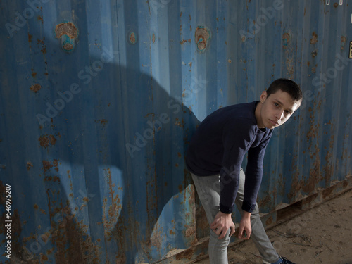 attractive and stylish fair-haired young caucasian guy posing with a serious face dressed in a sweater and jeans in an industrial area against a blue iron wall and a blue cloudy sky. lower angle