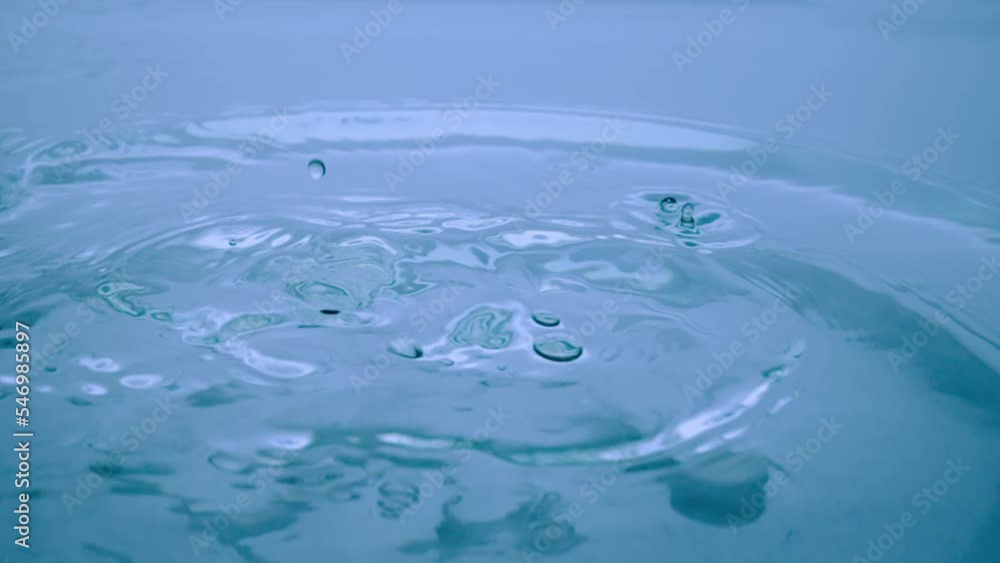 Macro shot of a drops of water falls on a blue liquid surface, creating water rings and waves. The dripping of a drop into the water and divergent circles on the water surface. Ripples on the surface. Macro shot of a drops of water falls on a blue liquid surface, creating water rings and waves. The dripping of a drop into the water and divergent circles on the water surface. Ripples on the surface.