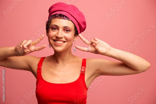Young athletic woman with a short haircut and purple hair in a red top and a pink hat with an athletic figure smiles and grimaces looking at the camera on a pink background © SHOTPRIME STUDIO