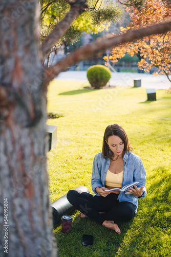 Attractive sporty woman sitting on grass under a tree at park, reading book in the park with bright sunlight. Full body size photo. Summer nature.