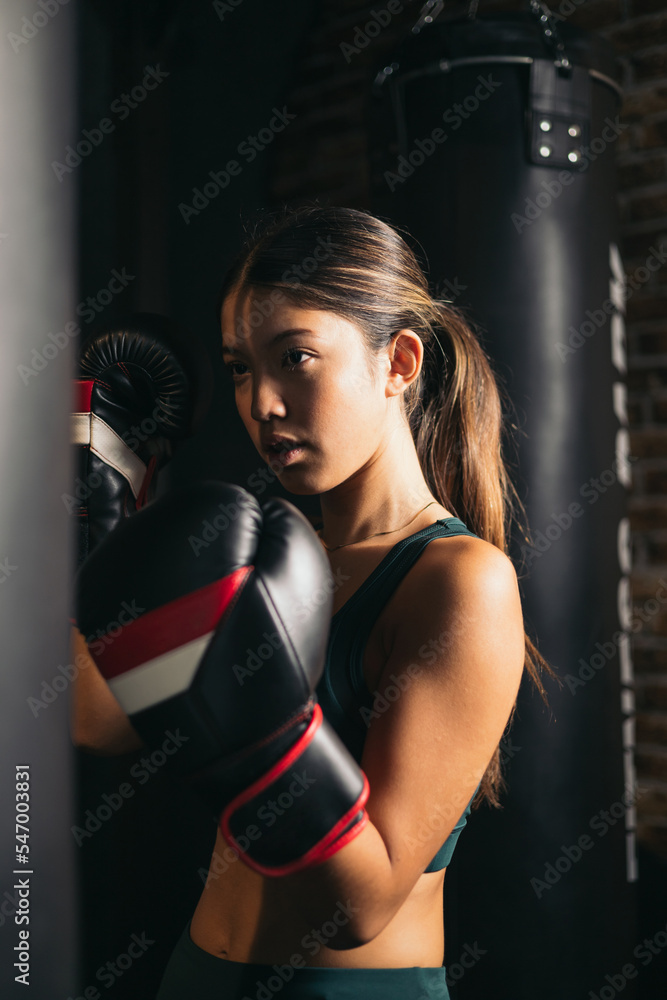 young asian woman in kickboxing guard position, training punching bag hard, boxing in gym class. Sport recreational activity, functional training, healthy lifestyle concept.