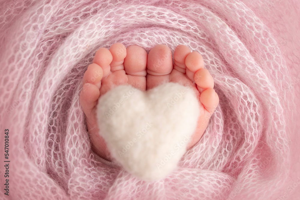 Knitted white heart in the legs of a baby. Soft feet of a new born in a pink wool blanket. Close-up of toes, heels and feet of a newborn. Macro photography the tiny foot of a newborn baby. Knitted white heart in the legs of a baby. Soft feet of a new born in a pink wool blanket. Close-up of toes, heels and feet of a newborn. Macro photography the tiny foot of a newborn baby.
