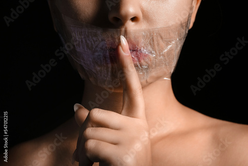 Young woman with taped mouth showing silence gesture on dark background, closeup. Censorship concept photo