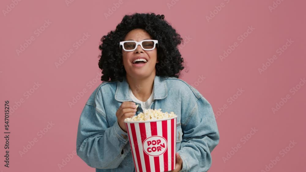 Portrait of laughing out loud African American young woman watching comedy movie or standup on TV with 3D glasses and eating popcorn. Positive woman laughing in studio on isolated pink background 4K Portrait of laughing out loud African American young woman watching comedy movie or standup on TV with 3D glasses and eating popcorn. Positive woman laughing in studio on isolated pink background 4K