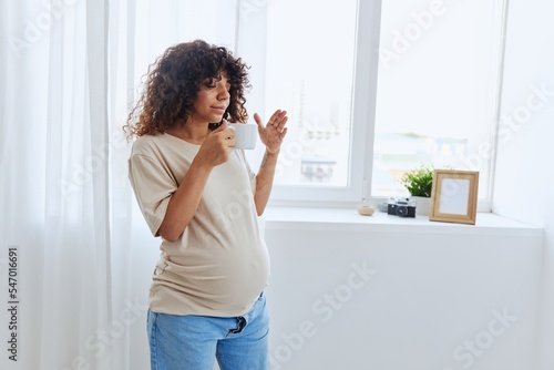 A pregnant woman stands at the window with a mug of warm water and tea and enjoys the view in a home t-shirt, the happiness of motherhood