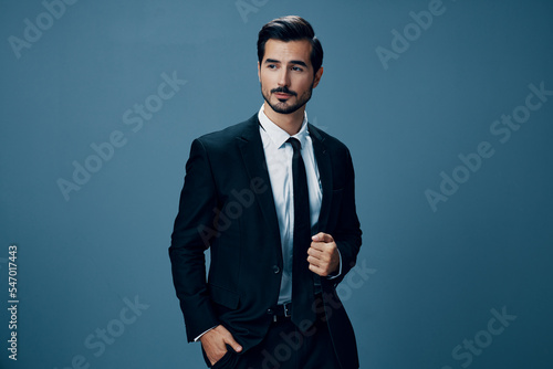 A confident business man smiles and looks at the camera in a black business suit on a blue background brunette with a beard