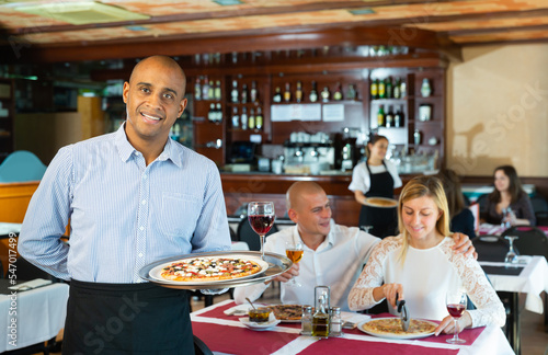 Smiling waiter holding serving tray with pizza at restaurant with customers his behind