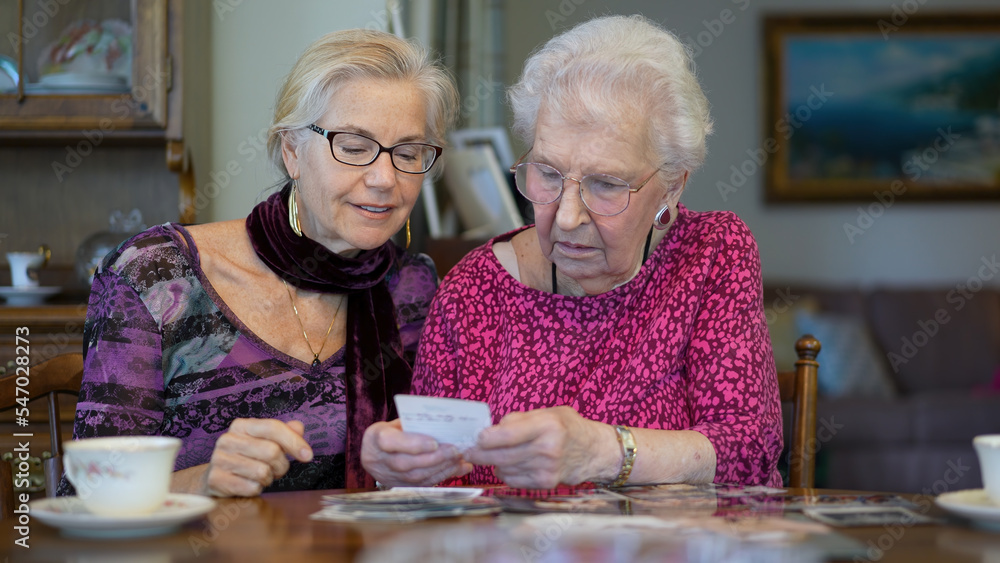 Senior elderly smiling happy woman looking at old photos having tea and remembering memories with daughter at the dining room table. Senior elderly smiling happy woman looking at old photos having tea and remembering memories with daughter at the dining room table.
