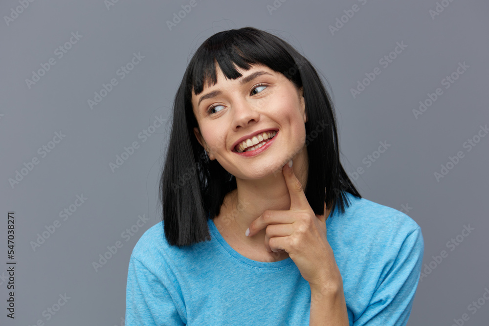 a beautiful, attractive, happy woman poses standing on a gray background in a blueT-shirt and happily looking at the camera with her index finger touching her face. horizontal studio photo a beautiful, attractive, happy woman poses standing on a gray background in a blueT-shirt and happily looking at the camera with her index finger touching her face. horizontal studio photo