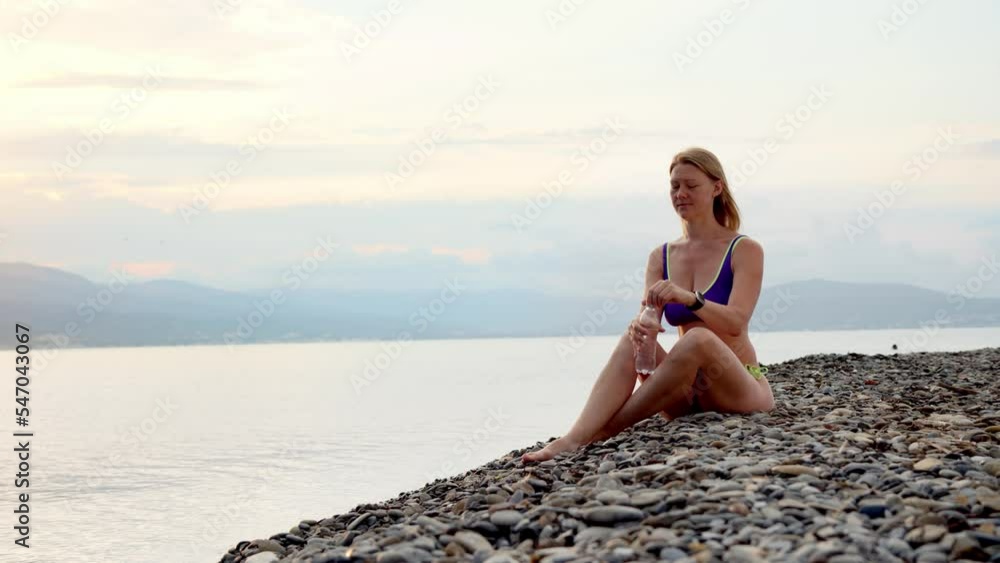 woman beautiful slender girl in purple bikini sits on shore of stone beach of ocean or sea with mountains background and drink water from transparent plastic bottle at sunset or dawn. woman beautiful slender girl in purple bikini sits on shore of stone beach of ocean or sea with mountains background and drink water from transparent plastic bottle at sunset or dawn.