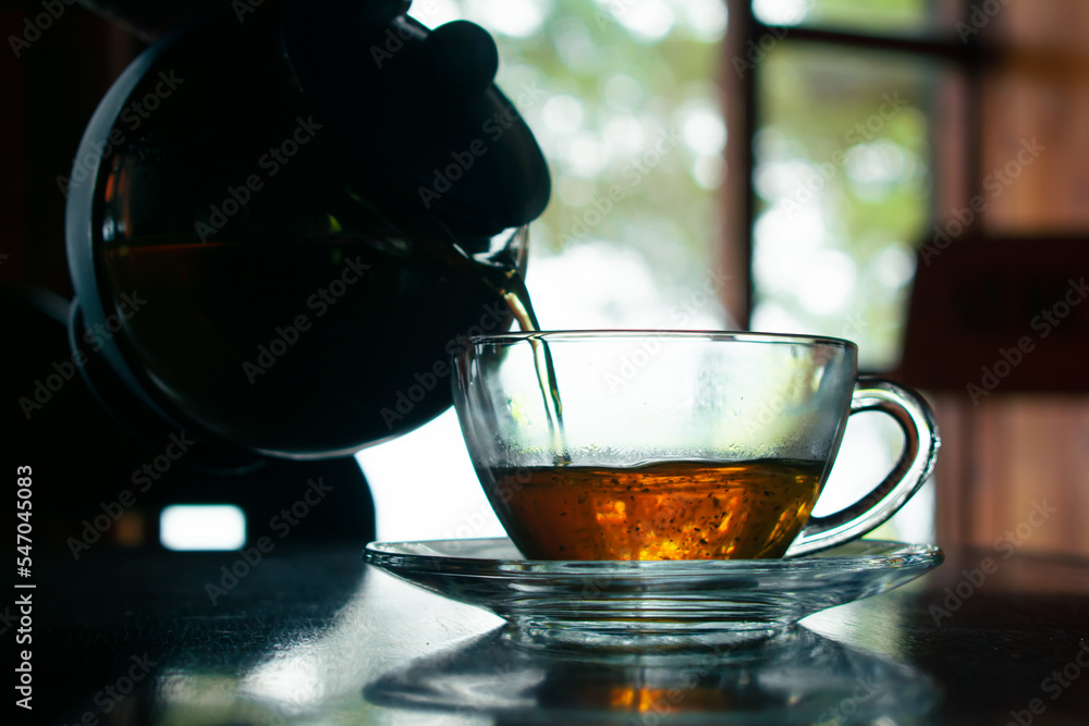 Pouring hot tea into a transparent cup by the window with a rustic blurred background.