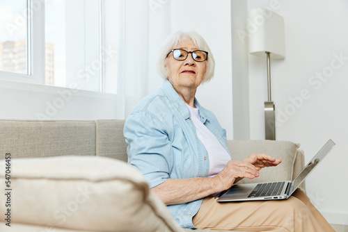 a cute elderly businesswoman is sitting on the sofa in a comfortable environment in stylish clothes and typing on a laptop while working from home holding it on her lap