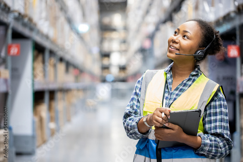Portrait of young attractive African American woman auditor or trainee staff work looking up stocktaking inventory in warehouse store by computer tablet and headphones near products shelf. Call center