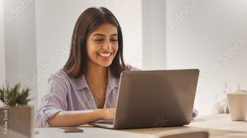 Internet, laptop and woman from India in home office, happy freelance worker at desk. Indian startup businesswoman with smile, technology and online project for trendy social media management company