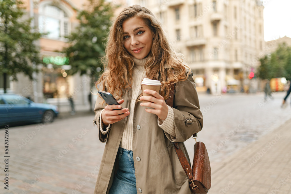Young woman with curly blonde hair using the phone with a cup of coffee in hands on the city streets Young woman with curly blonde hair using the phone with a cup of coffee in hands on the city streets
