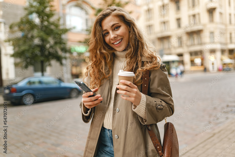Young woman with curly blonde hair using the phone with a cup of coffee in hands on the city streets Young woman with curly blonde hair using the phone with a cup of coffee in hands on the city streets