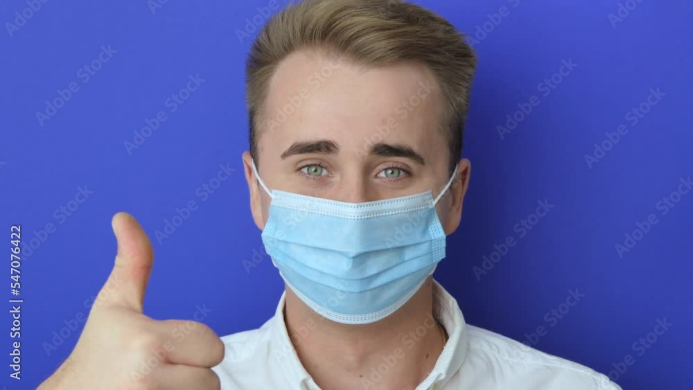 Closeup shot of a young man using a medical mask during the Covid-19 pandemic. Guy wears a surgical mask and shows a thumbs-up sign to protect himself from coronavirus Closeup shot of a young man using a medical mask during the Covid-19 pandemic. Guy wears a surgical mask and shows a thumbs-up sign to protect himself from coronavirus