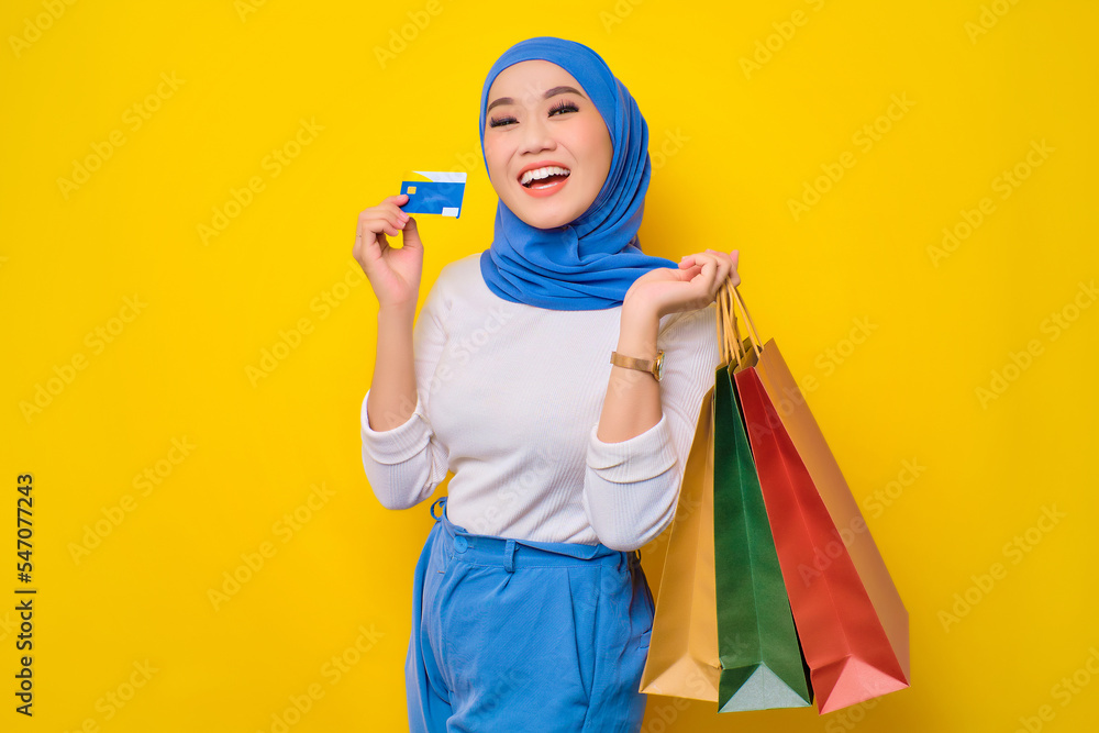 Cheerful young Asian Muslim woman holding credit card and shopping bags, looking at camera isolated on yellow background Cheerful young Asian Muslim woman holding credit card and shopping bags, looking at camera isolated on yellow background