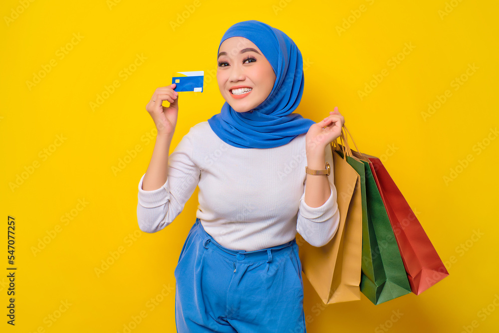 Cheerful young Asian Muslim woman holding credit card and shopping bags, looking at camera isolated on yellow background Cheerful young Asian Muslim woman holding credit card and shopping bags, looking at camera isolated on yellow background