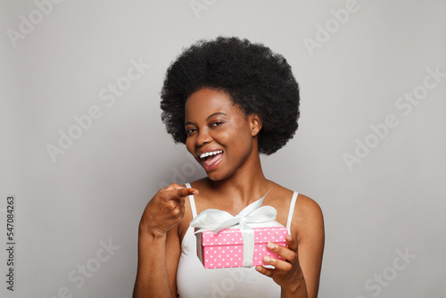 Cute cheerful woman smiling and pointing finger at pink gift present box against white studio wall background