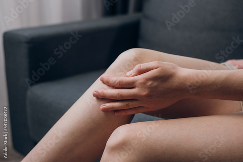Canvas Print Closeup of unhealthy young woman sitting on sofa and feeling knee pain, doing massage knee at home