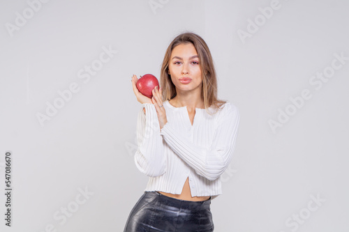 A beautiful young brown-haired woman holds a red apple in her hands. The girl is dressed in a knitted blouse and black leather pants on a white background