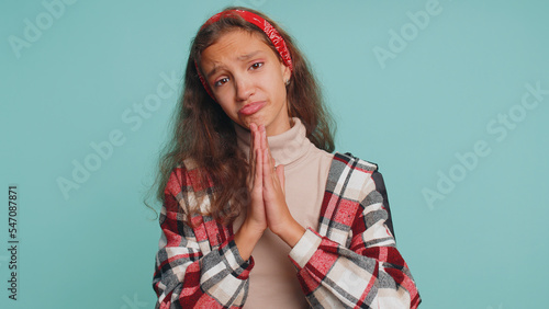 Please, God help. Young teenager child girl kid sincerely praying to God and looking up with pleading expression, begging apology, beseeching with hopeful face. Preteen children on blue background © Andrii Iemelianenko