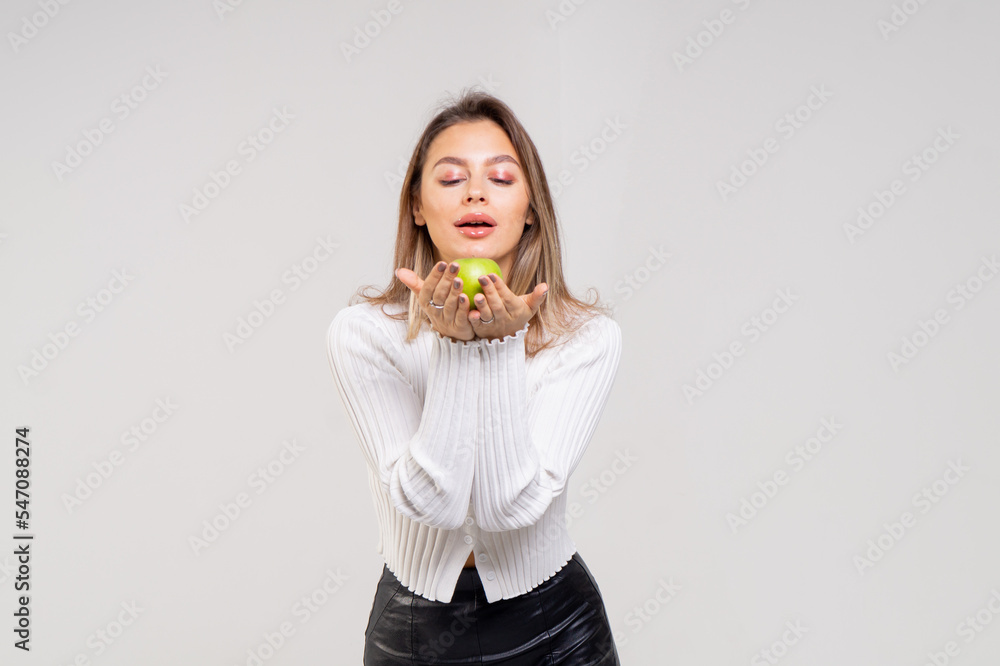 Cute young brown-haired woman posing with a yellow apple on a white background. The girl is dressed in a white knitted blouse and black leather pants