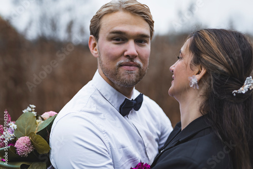 Proud groom - 30-year-old bearded blond Scandinavian man - looking at camera with a soft smile on his face while his beloved spouse is cuddling him. Blurred background. High quality photo © PoppyPix